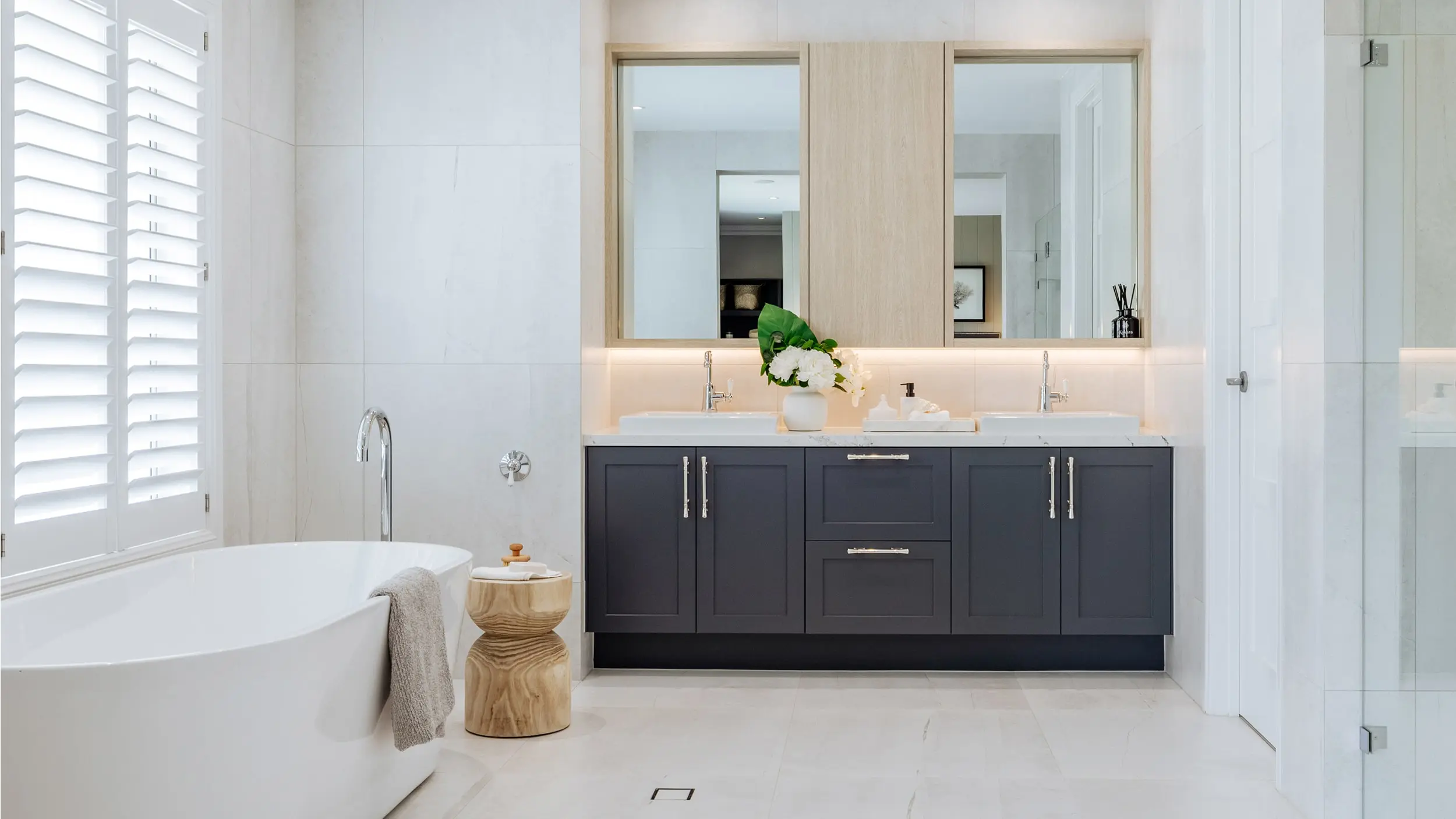 Modern bathroom with white freestanding tub, navy blue double vanity, white countertops, and wooden accents.