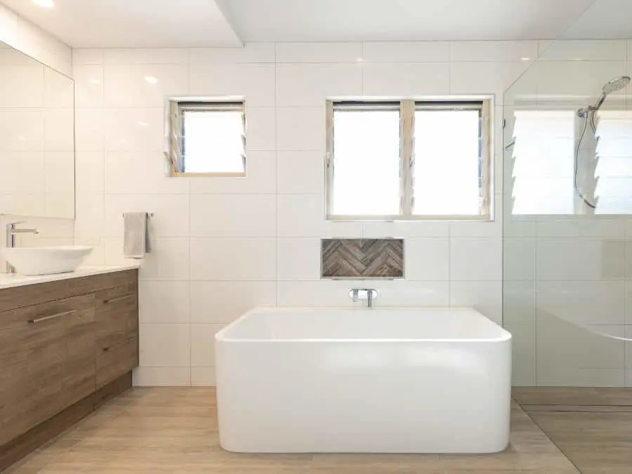 Modern white bathroom with freestanding tub, wooden vanity, glass shower, and windows with herringbone tile accent.