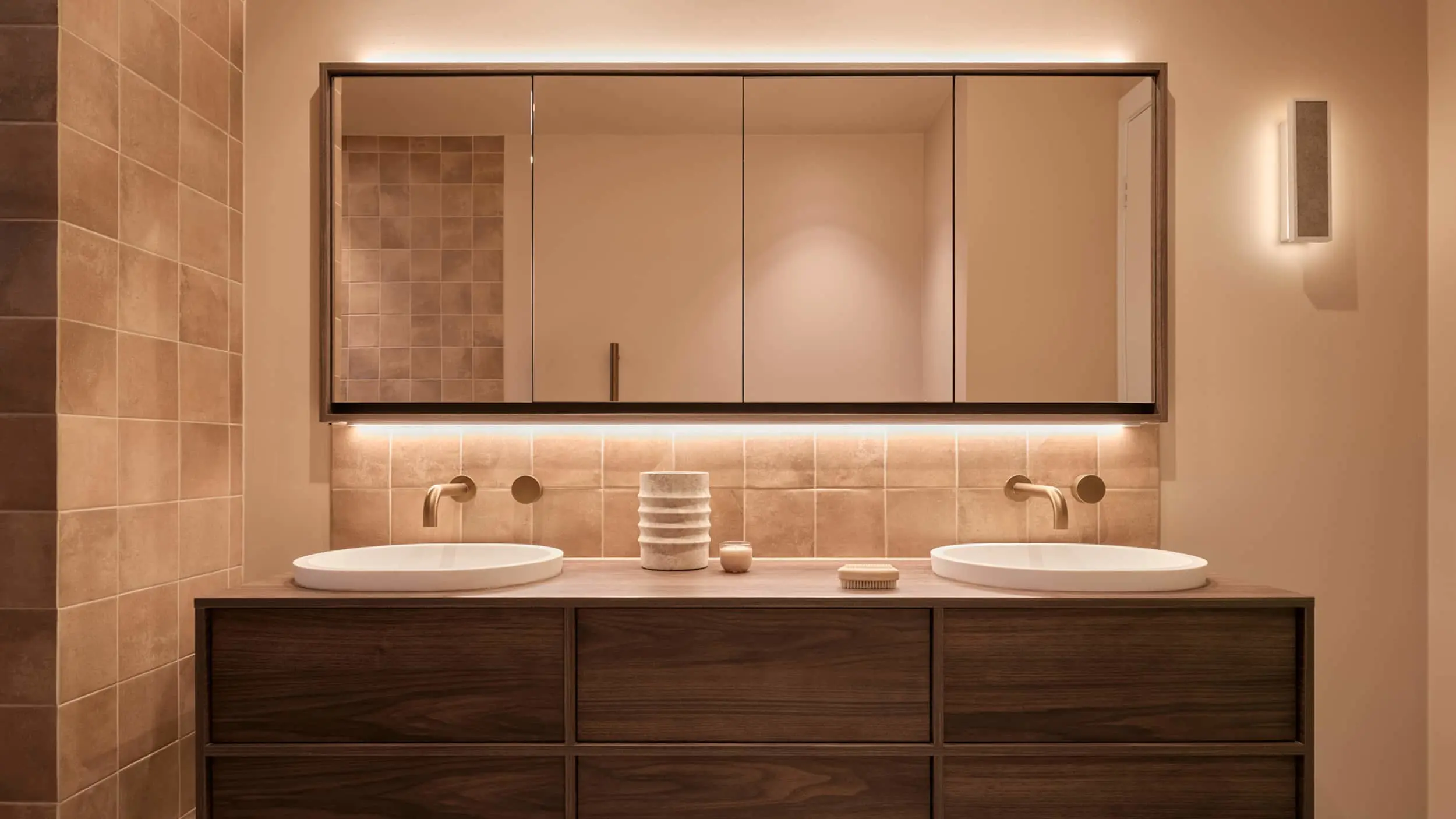 Modern bathroom with double sinks, wooden vanity, mirrored cabinets, and warm lighting against beige tiled walls.