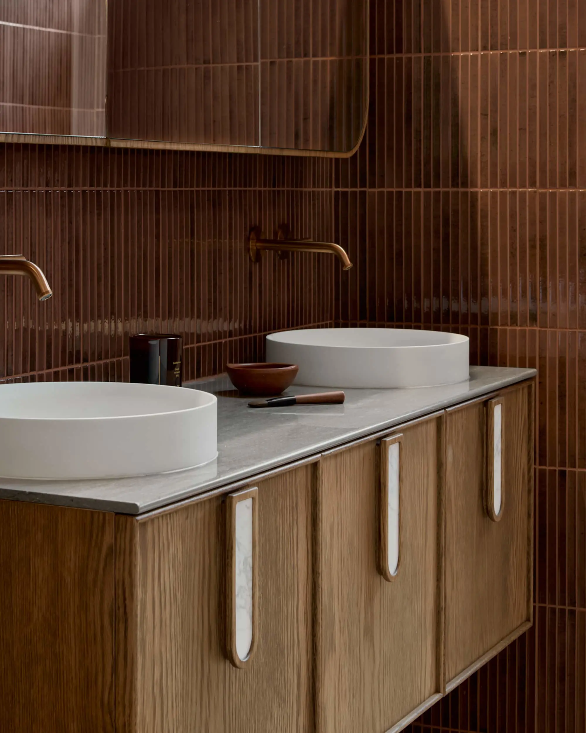 Modern bathroom with wooden vanity, white vessel sinks, brass wall-mounted faucets, and vertical brown tile backsplash.
