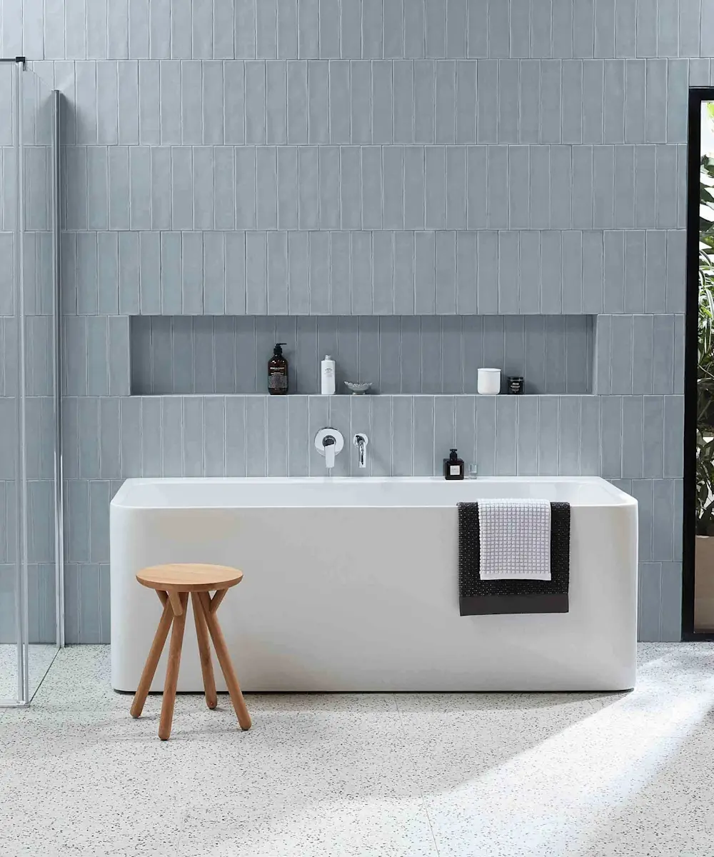 Modern bathroom with white freestanding bathtub, wooden stool, gray tiled walls, and built-in shelf with toiletries.