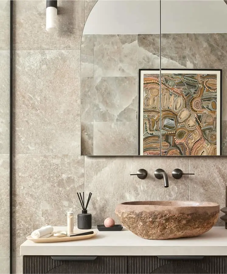 Modern bathroom with stone vessel sink, beige marble walls, indigenous artwork, and minimalist accessories on white countertop.