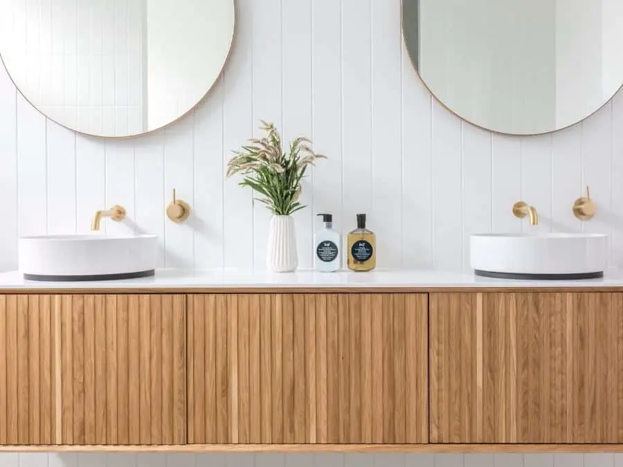 Modern bathroom vanity with wooden cabinets, white vessel sinks, round mirrors, brass fixtures, and decorative plant.
