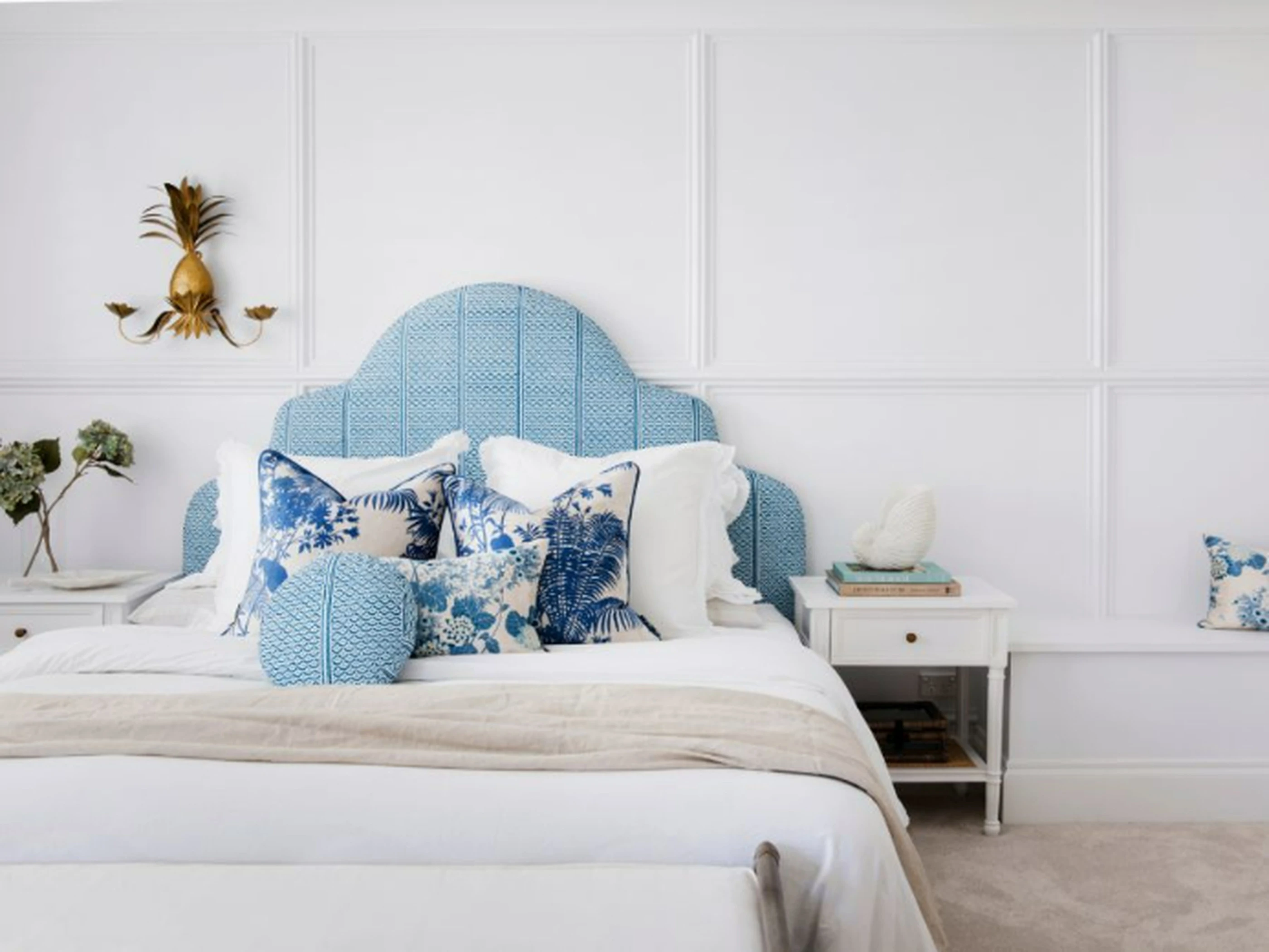 Bedroom with blue upholstered headboard, white bedding, blue floral pillows, and gold pineapple wall sconce.