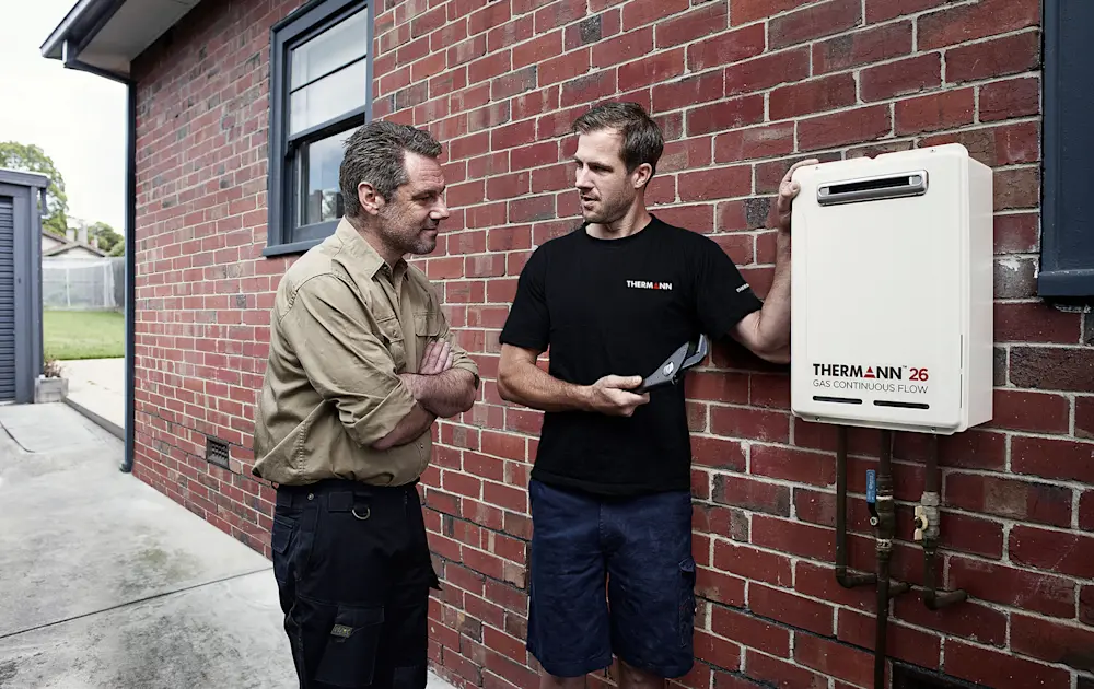 Two men discussing a Thermann 26 gas continuous flow unit mounted on a brick wall outside a home.