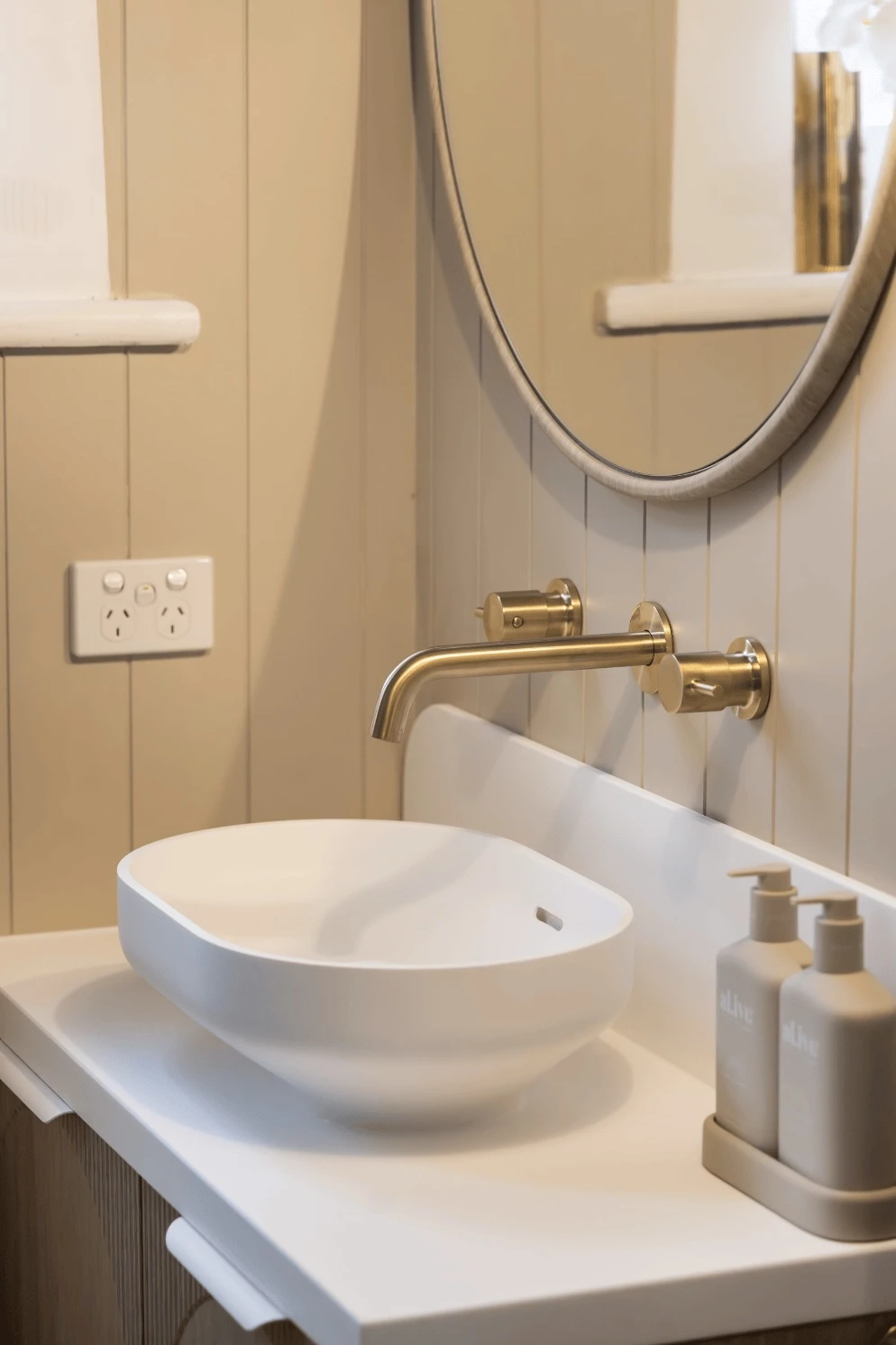 Modern bathroom with white vessel sink, brass wall-mounted faucet, round mirror, and neutral-colored soap dispensers.