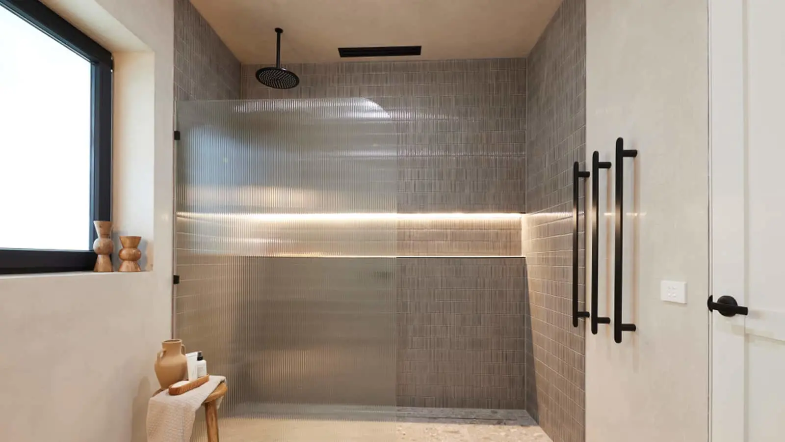 Modern bathroom with glass shower enclosure, gray tiled walls, black rainfall showerhead, and wooden accent stool.