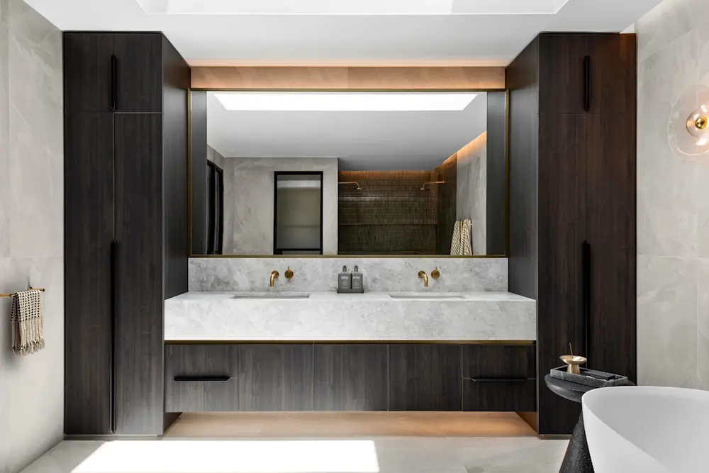 Modern bathroom with dark wood cabinetry, white marble countertop, gold fixtures, and large mirror with ambient lighting.
