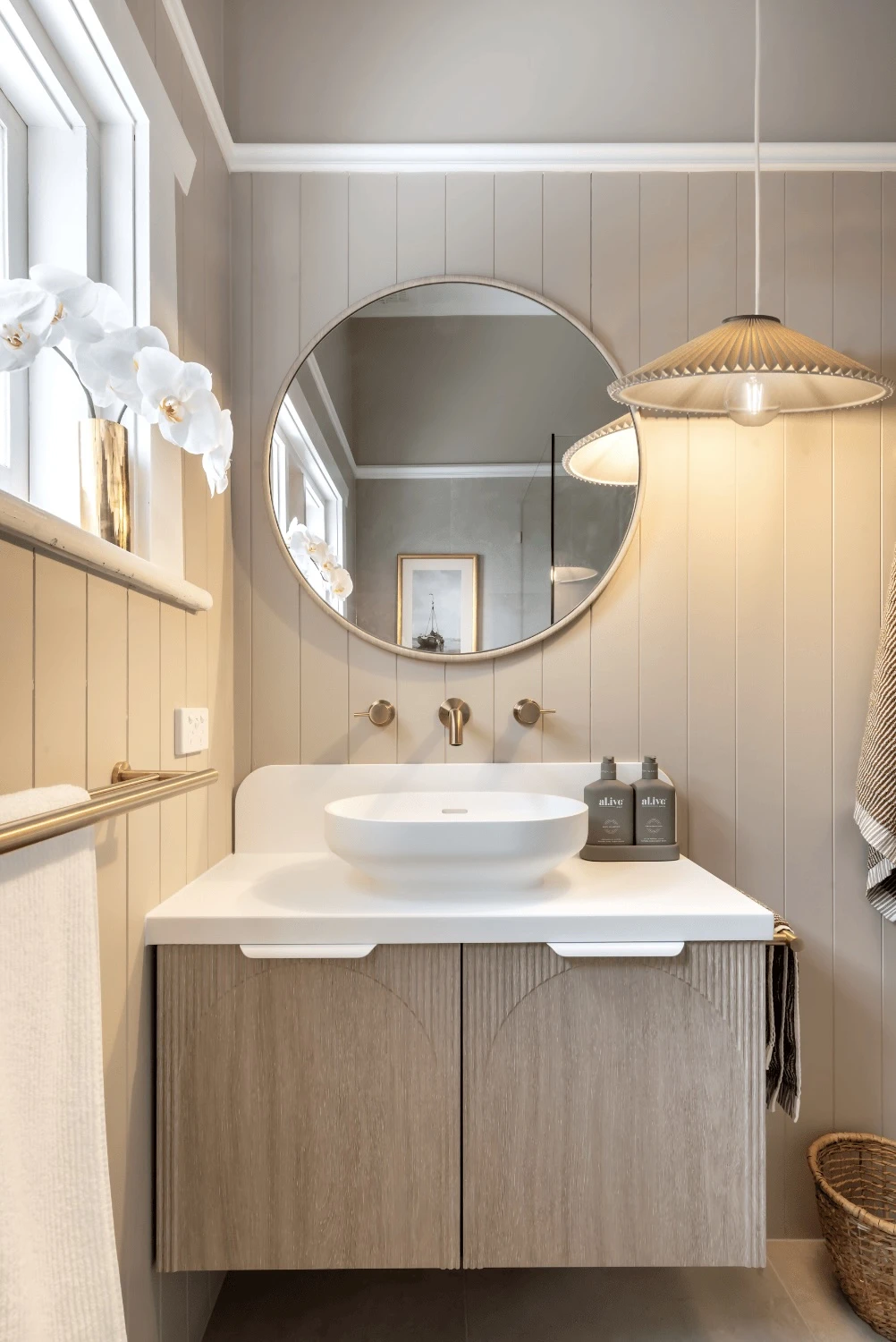Elegant bathroom with round mirror, white vessel sink, wooden vanity, pendant light, and white orchids in gold vase.