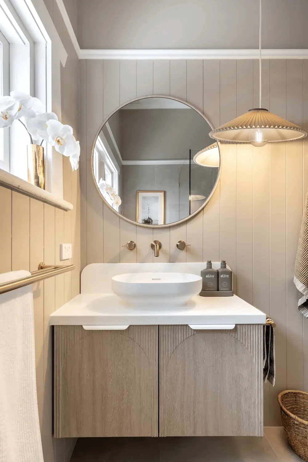 Elegant bathroom with round mirror, white vessel sink, wooden vanity, pendant light, and white orchids in gold vase.