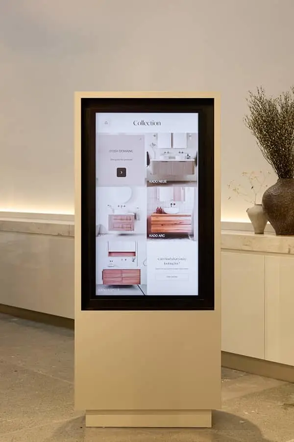 A freestanding digital kiosk displaying a bathroom collection catalogue, placed in a showroom with a vase nearby.