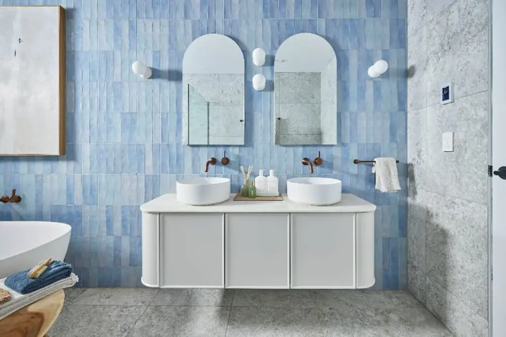 Modern bathroom with light blue tiled walls, white double vanity with vessel sinks, and arched mirrors with brass fixtures.