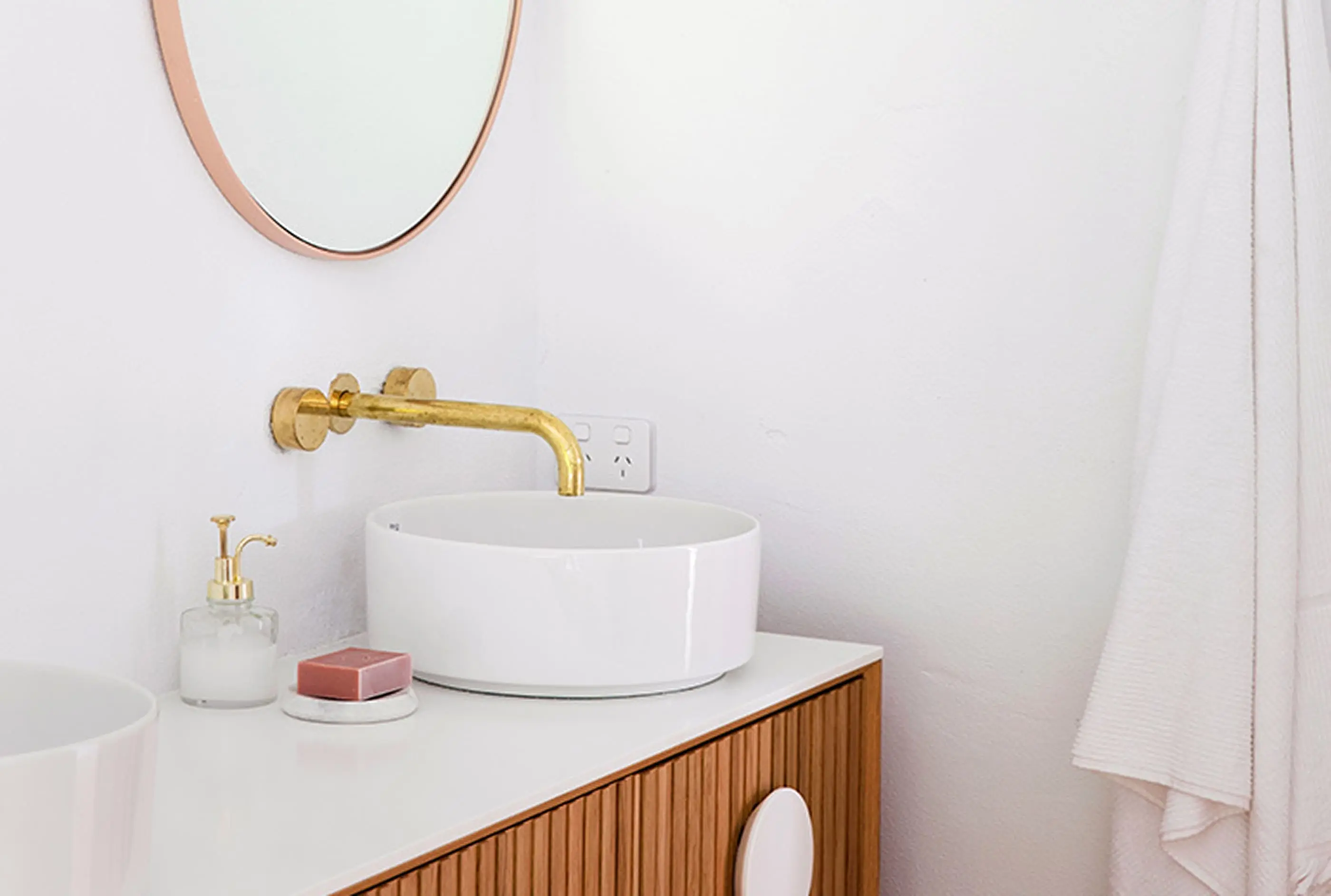 Modern bathroom with white vessel sink, brass wall-mounted faucet, wooden vanity, and round copper-framed mirror.