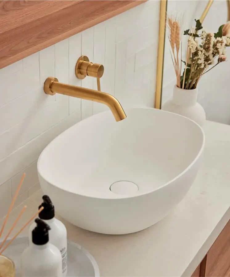 Modern bathroom sink with brass wall-mounted faucet, white vessel basin, and dried flowers in a vase nearby.