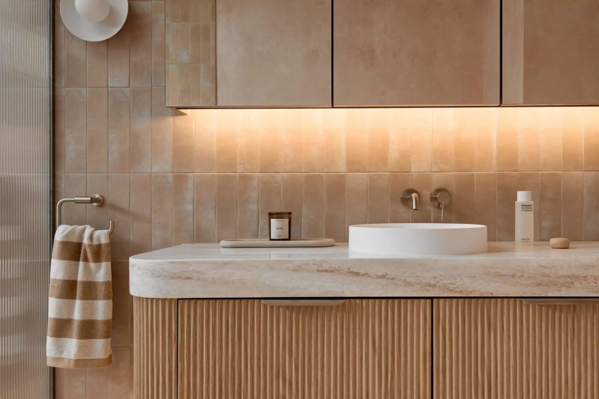Modern bathroom with beige tiled walls, marble countertop, white vessel sink, and wooden cabinetry with striped towel.