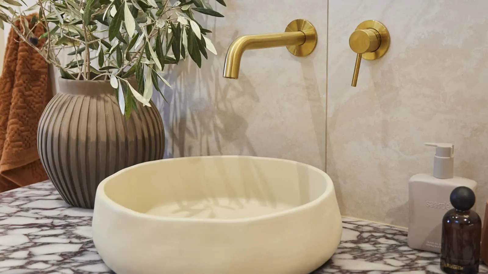 Modern bathroom with cream vessel sink, brass wall-mounted faucet, gray ribbed vase with olive branches on marble counter.