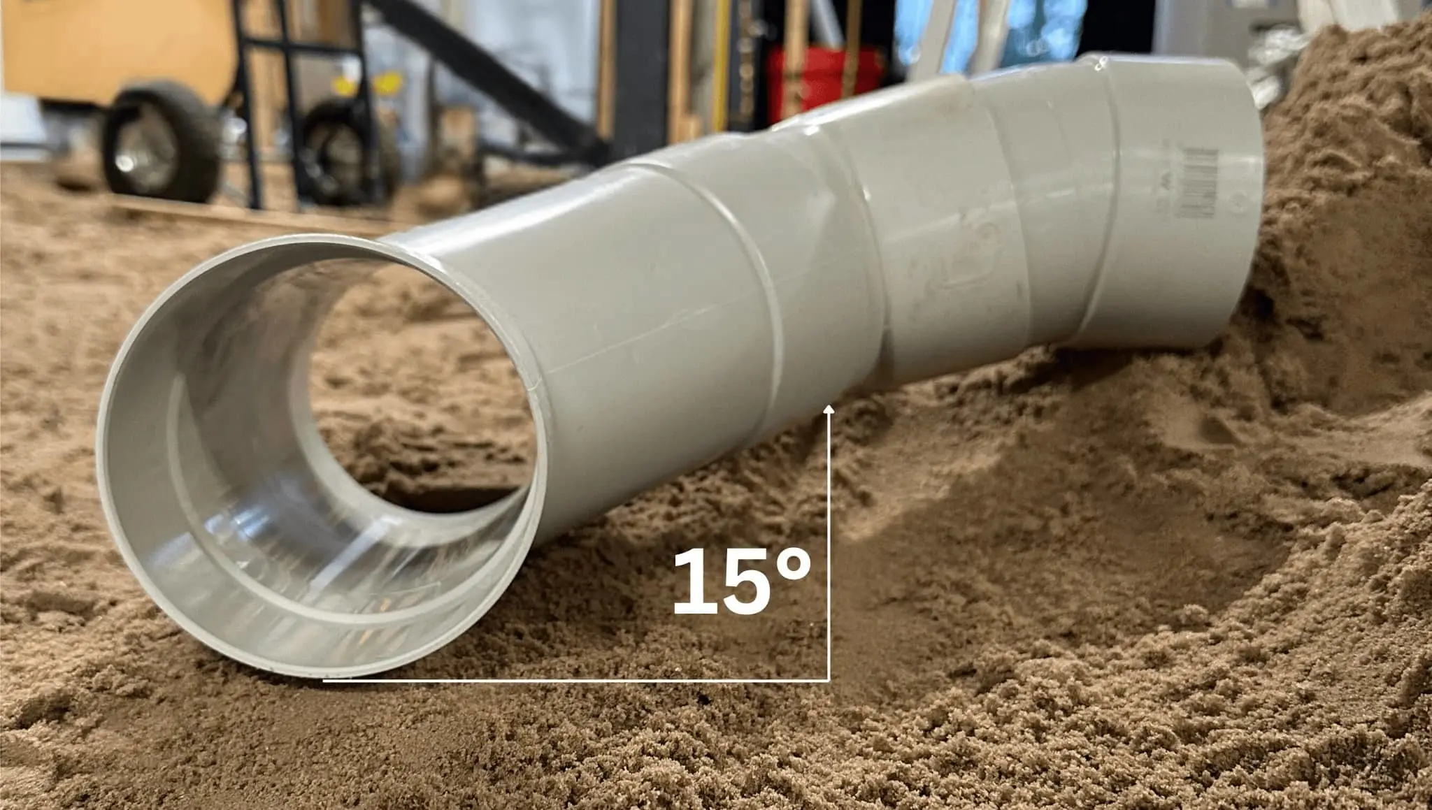 White PVC pipe with 15-degree angle bend partially buried in sand at a construction site.
