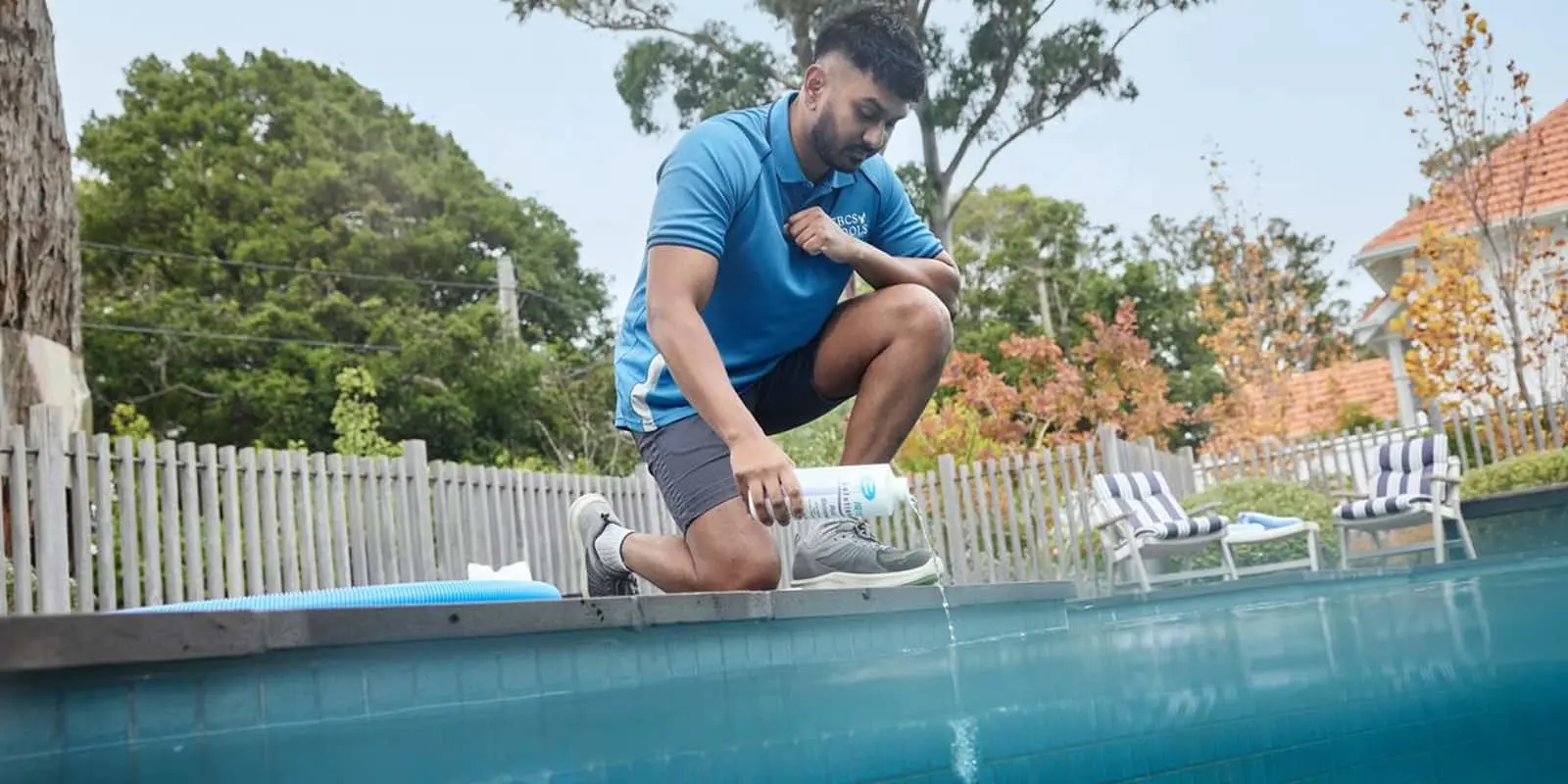 Person in blue polo shirt adding pool chemicals while kneeling at the edge of a swimming pool in a backyard setting.