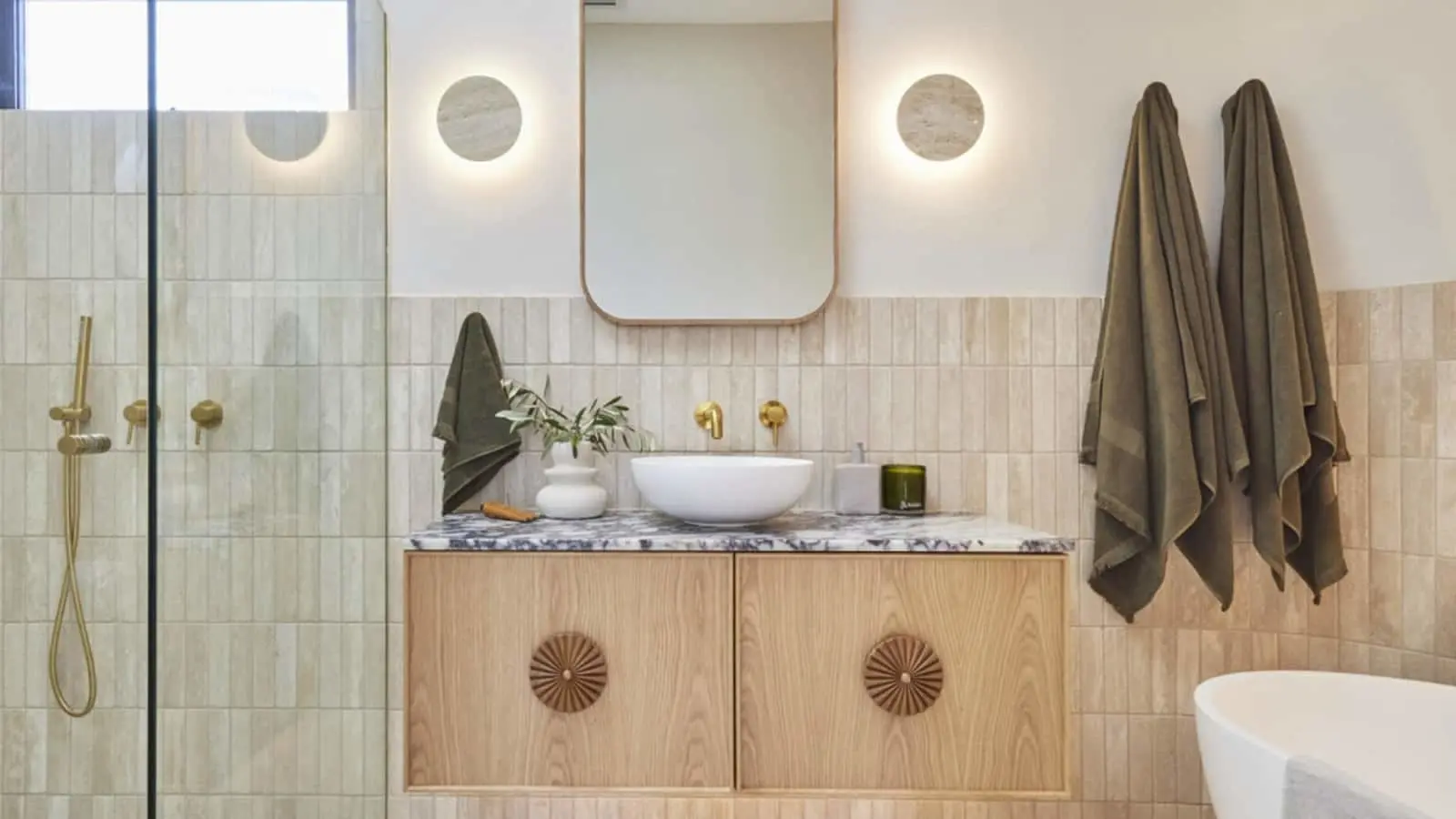 Modern bathroom with wooden vanity, white vessel sink, circular wall lights, olive towels, and glass shower enclosure.