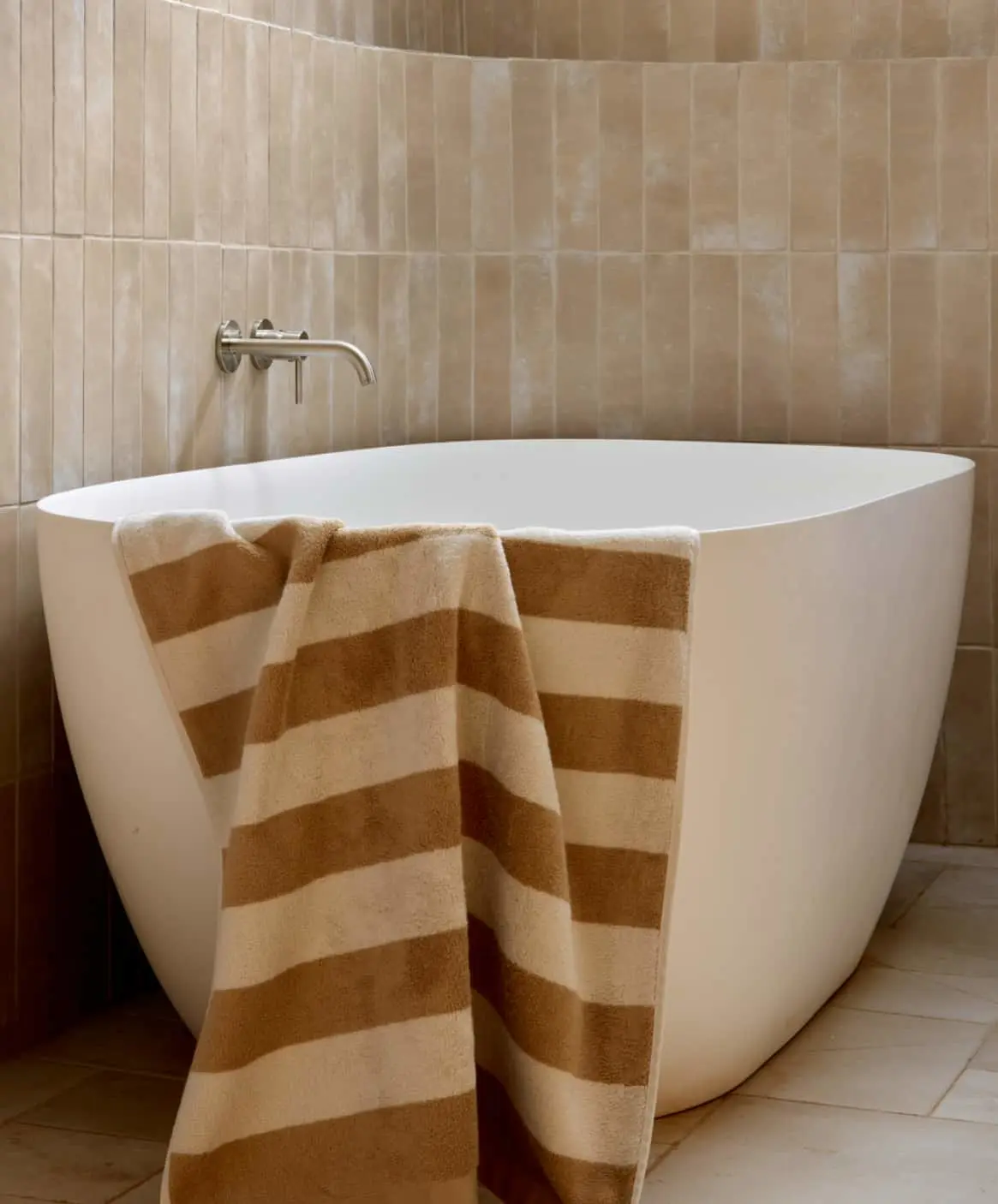 Modern bathroom with beige tiled walls, white freestanding tub, and brown striped towels.