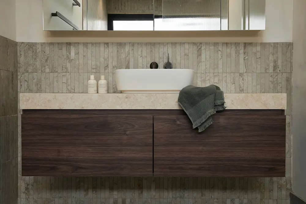 Modern bathroom vanity with white vessel sink, dark wood cabinets, stone countertop, and green towel on marble-tiled wall.