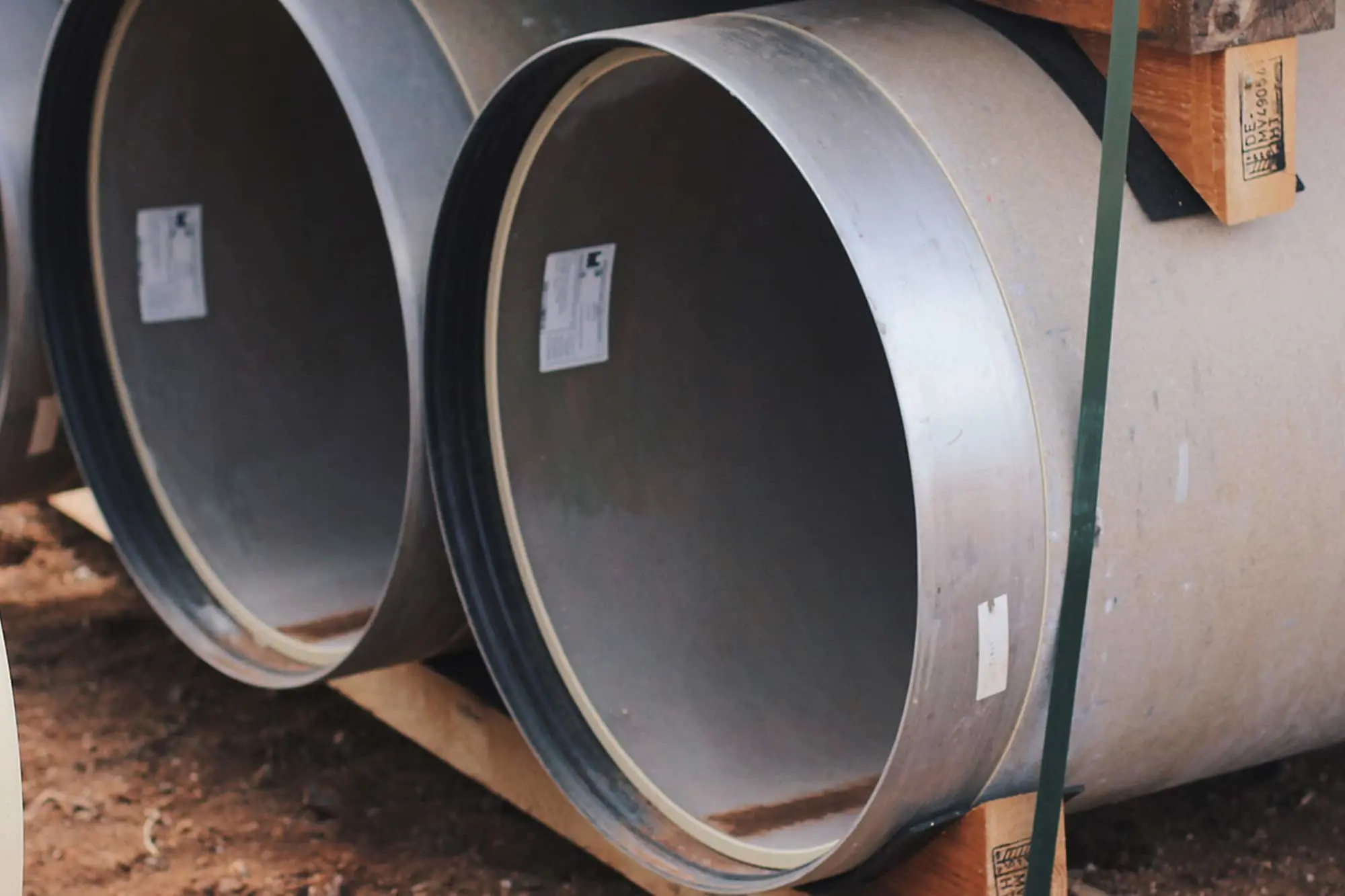 Two large metal pipes or culverts with labels attached, lying horizontally on wooden supports at a construction site.