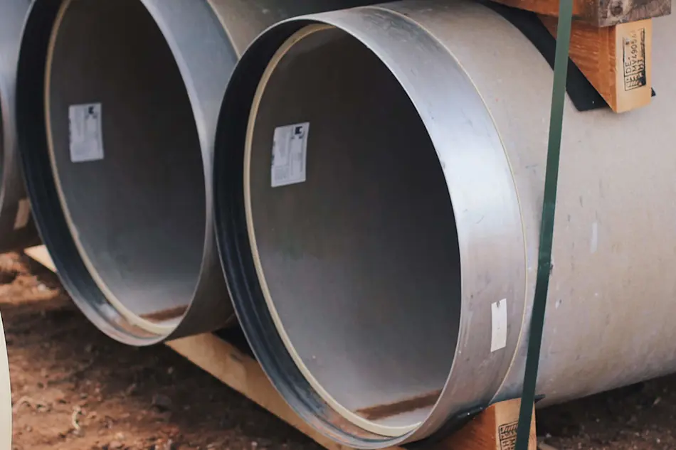 Two large metal pipes or culverts with labels attached, lying horizontally on wooden supports at a construction site.