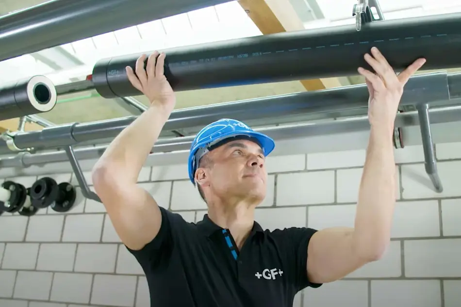 Worker in blue hard hat installing black pipe overhead in industrial setting with white tiled wall background.