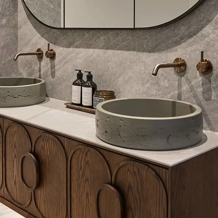 Modern bathroom with double concrete vessel sinks, wooden vanity, brass wall-mounted faucets, and curved mirror.