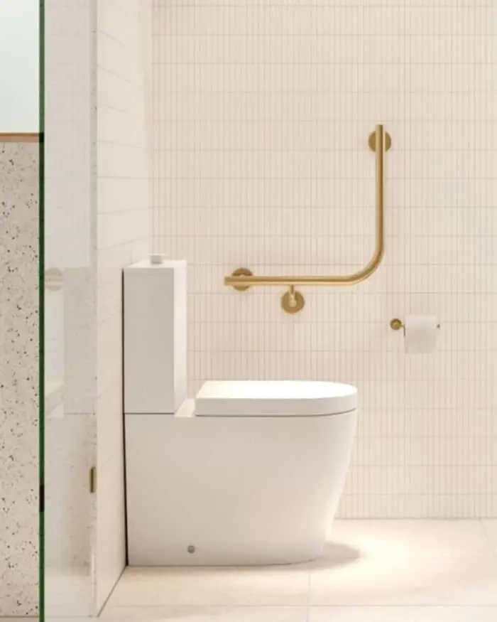 Modern bathroom with white toilet, brass grab bar, and cream tiled walls featuring accessible design elements.