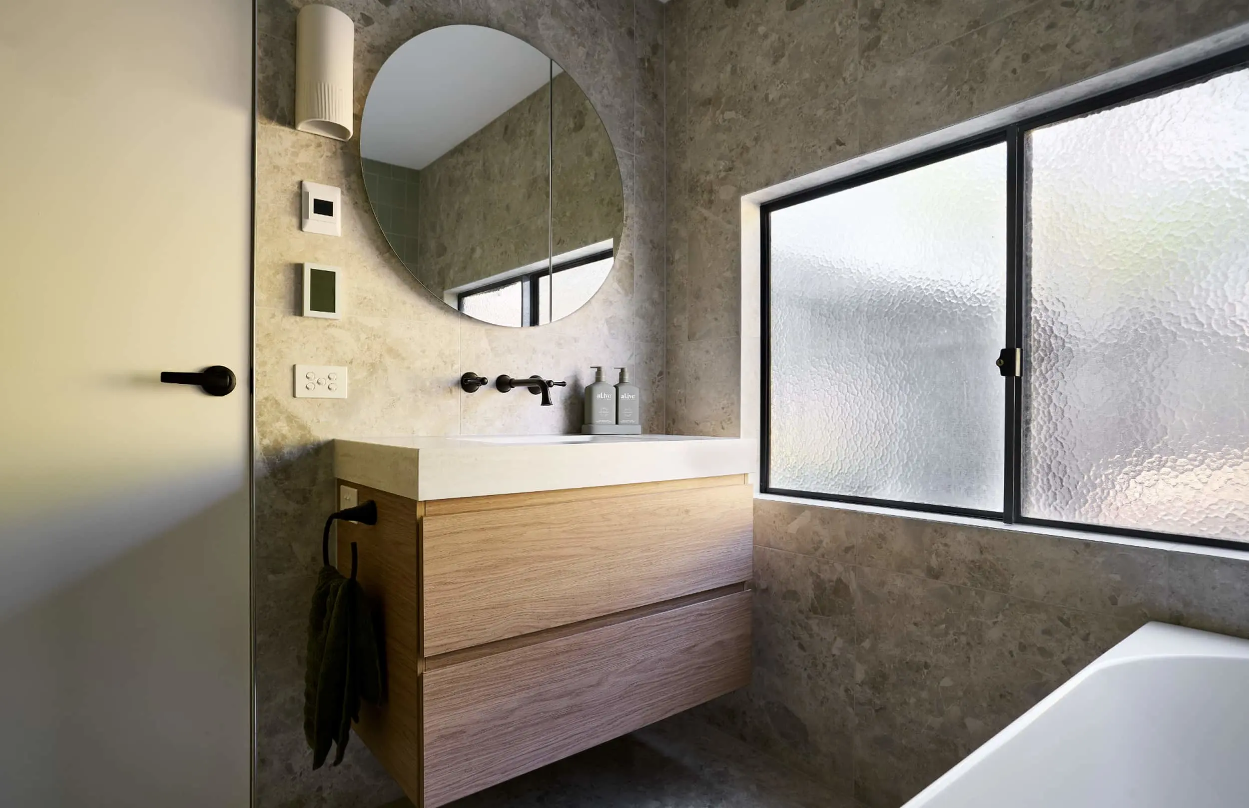Modern bathroom with concrete walls, wooden vanity, round mirror, black fixtures, and frosted window.