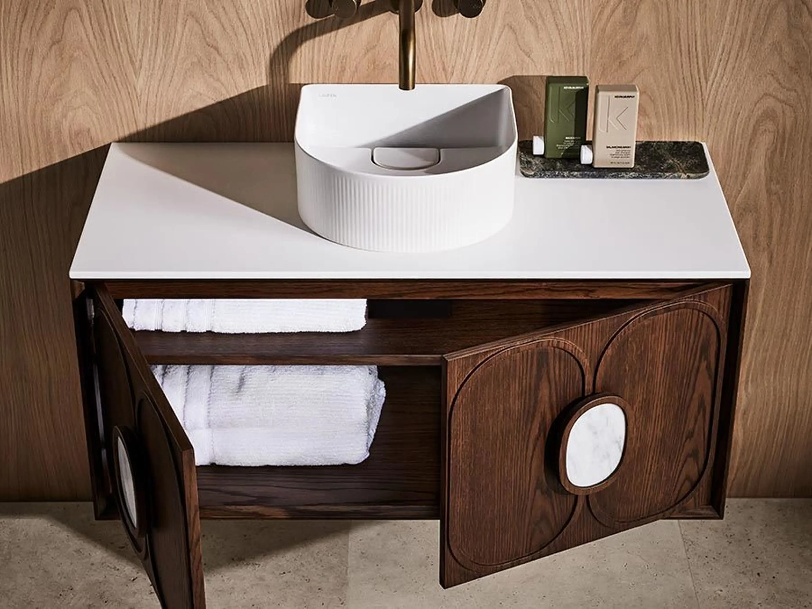 Modern bathroom vanity with white circular sink on wooden cabinet, brass faucet, and folded white towels inside.