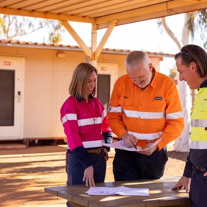 Three workers in high-visibility workwear reviewing documents at a wooden table under a covered outdoor area.