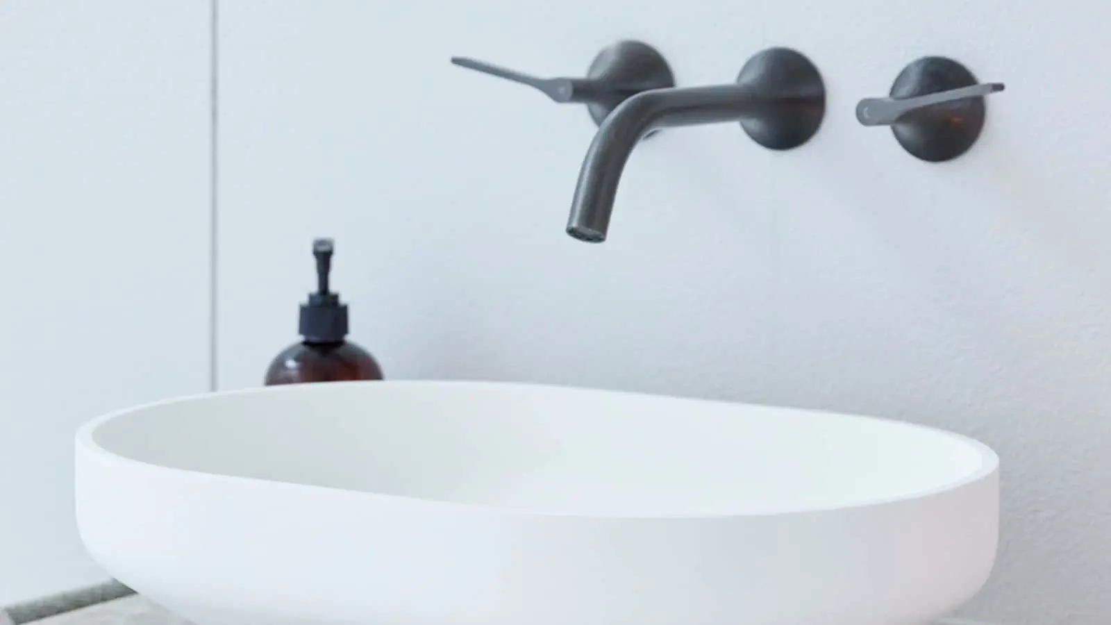 Modern white bathroom sink with wall-mounted black faucet and soap dispenser against light gray wall.