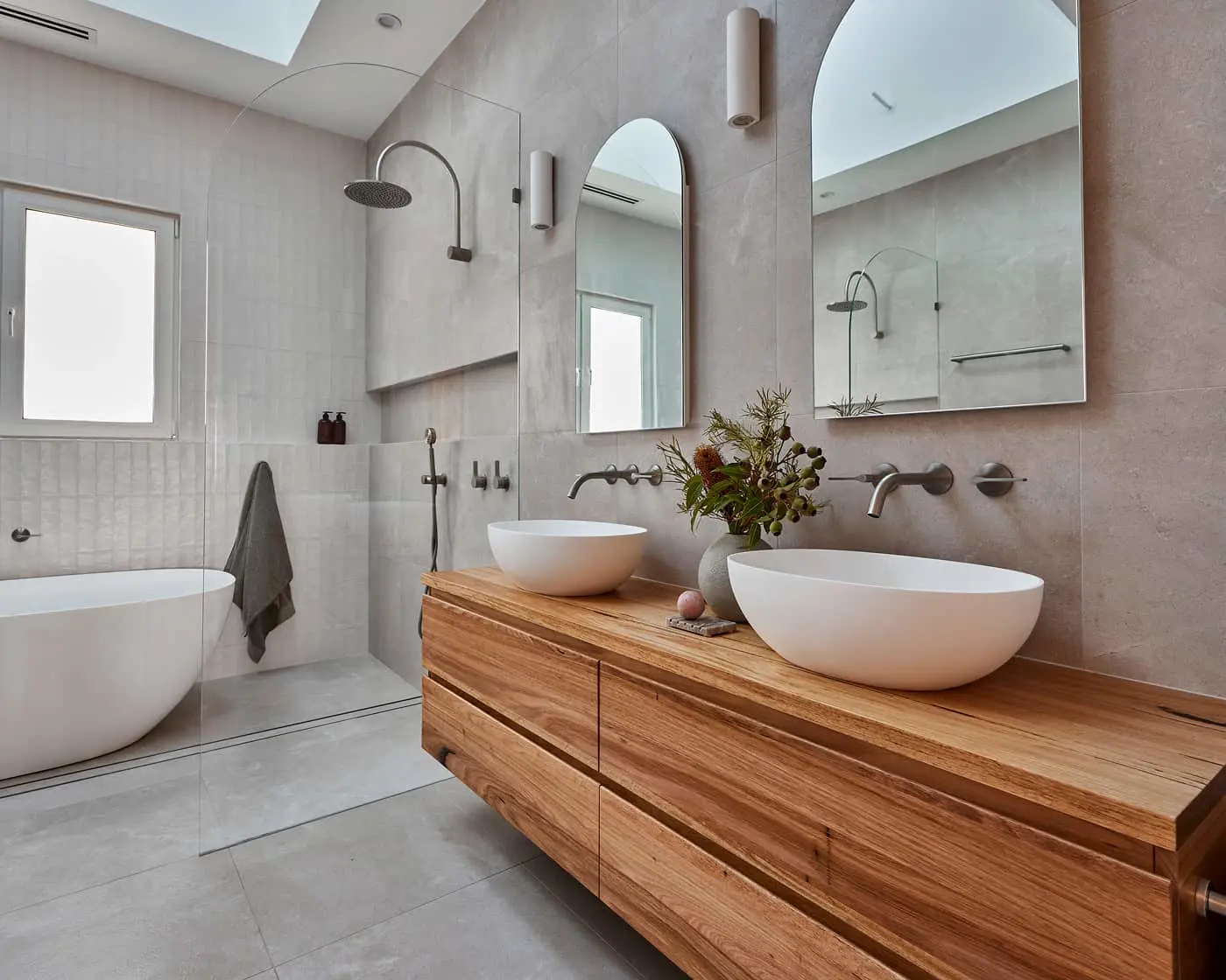 Modern bathroom with light gray tiles, wooden vanity with white vessel sinks, and a bathtub with rainfall shower.