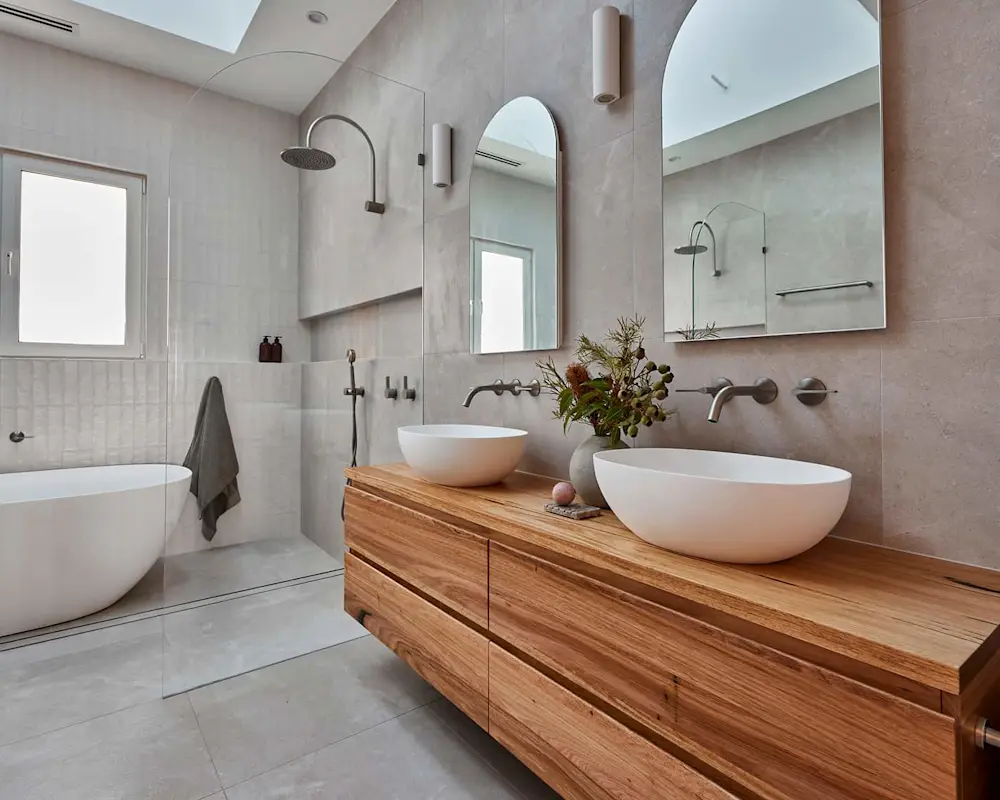 Modern bathroom with light gray tiles, wooden vanity with white vessel sinks, and a bathtub with rainfall shower.