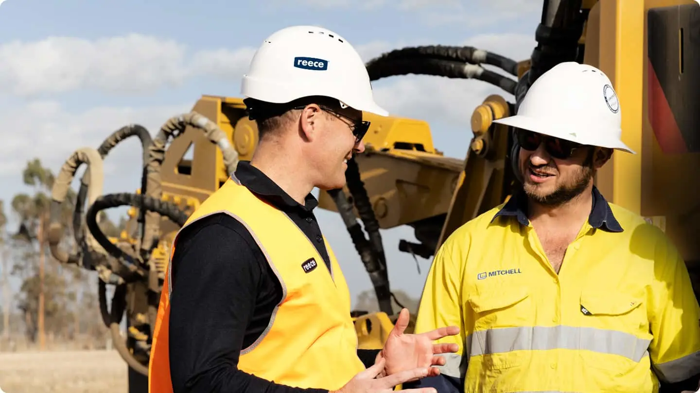 Two construction workers in white hard hats and high-visibility gear conversing near yellow heavy machinery.