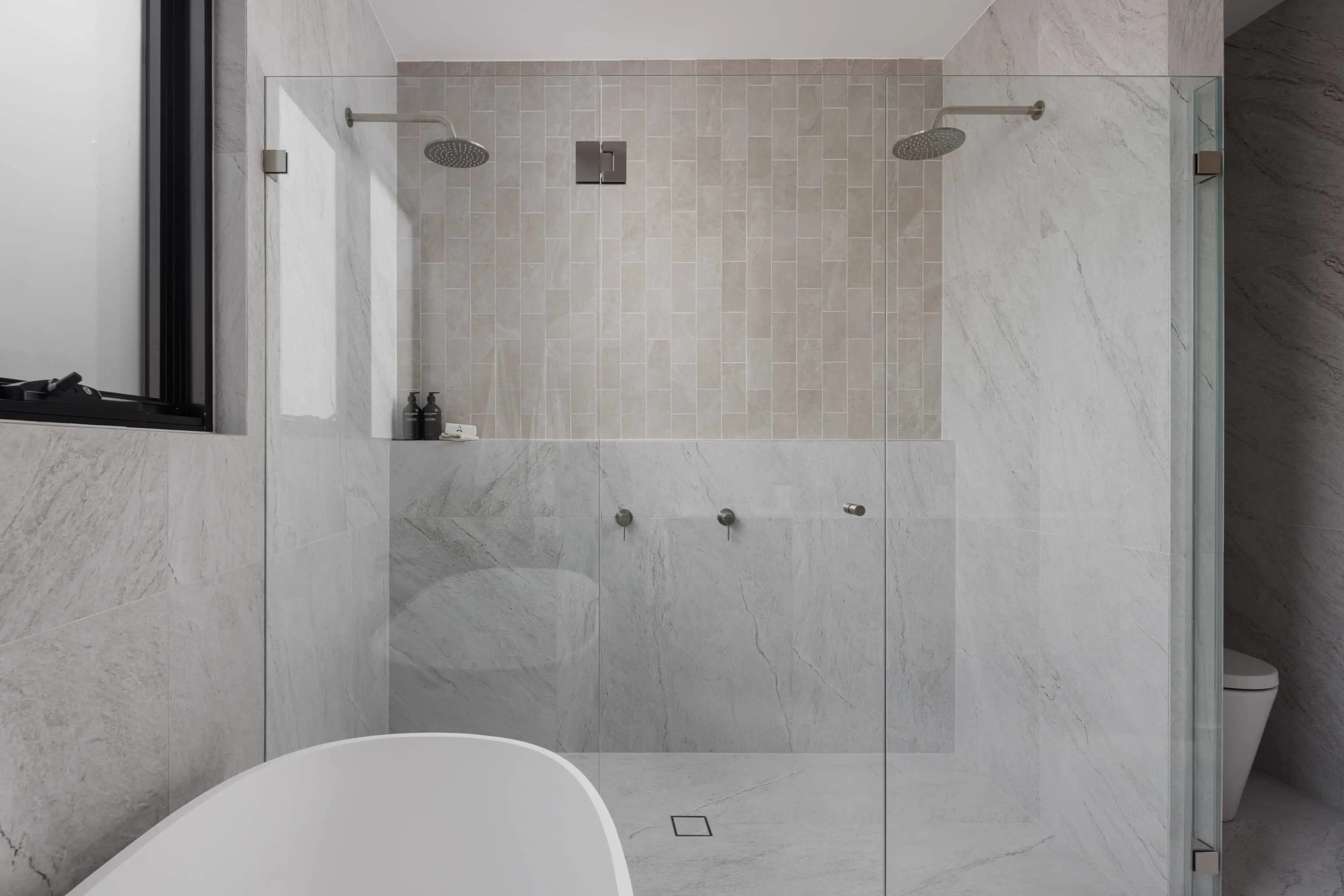 Modern bathroom with white marble walls, dual rainfall showerheads, beige tiled accent wall, and partial view of a freestanding tub.