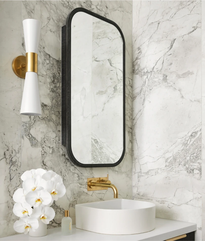 Modern bathroom with marble walls, black-framed mirror, white vessel sink, brass faucet, gold wall sconce, and white orchids.