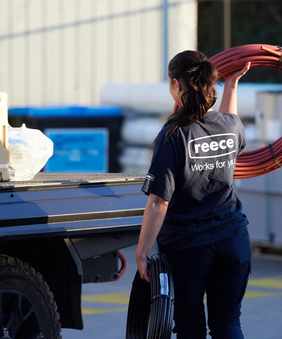 Worker in Reece shirt loading red coiled pipes onto a truck bed in an industrial setting.