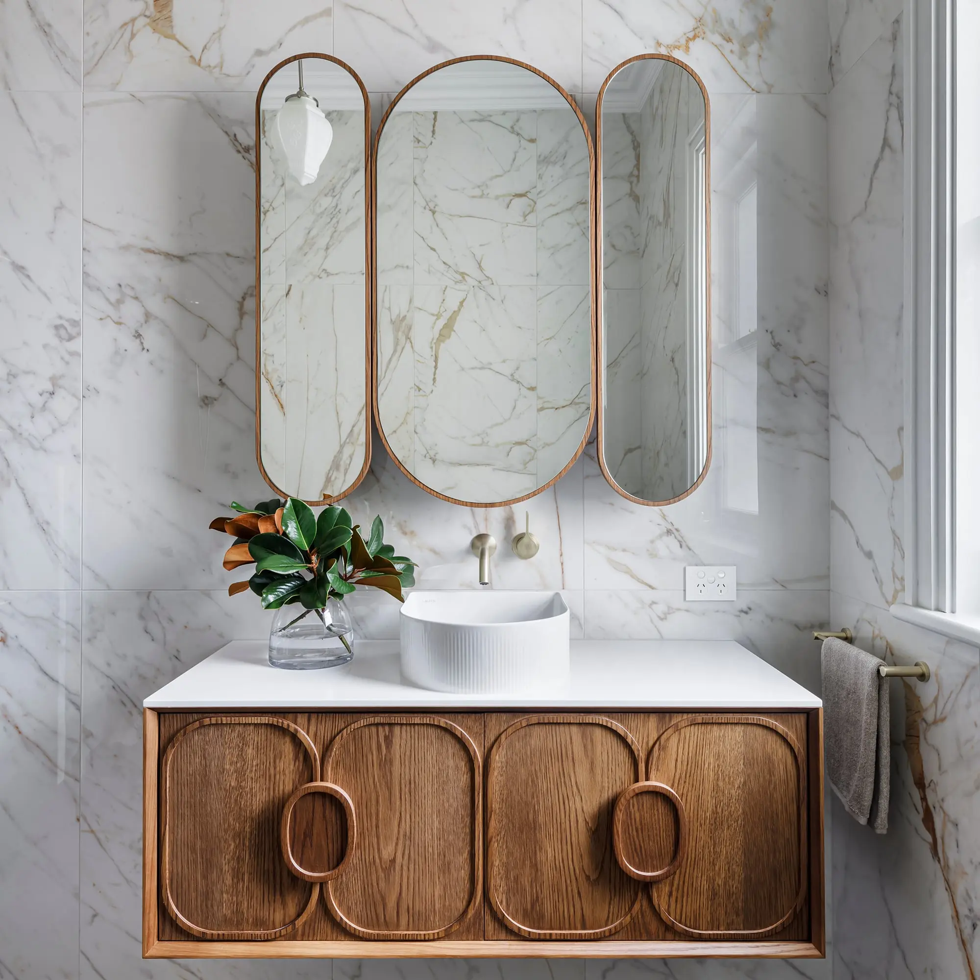 Modern bathroom vanity with wooden cabinet, white countertop, vessel sink, and round mirrors against marble walls.