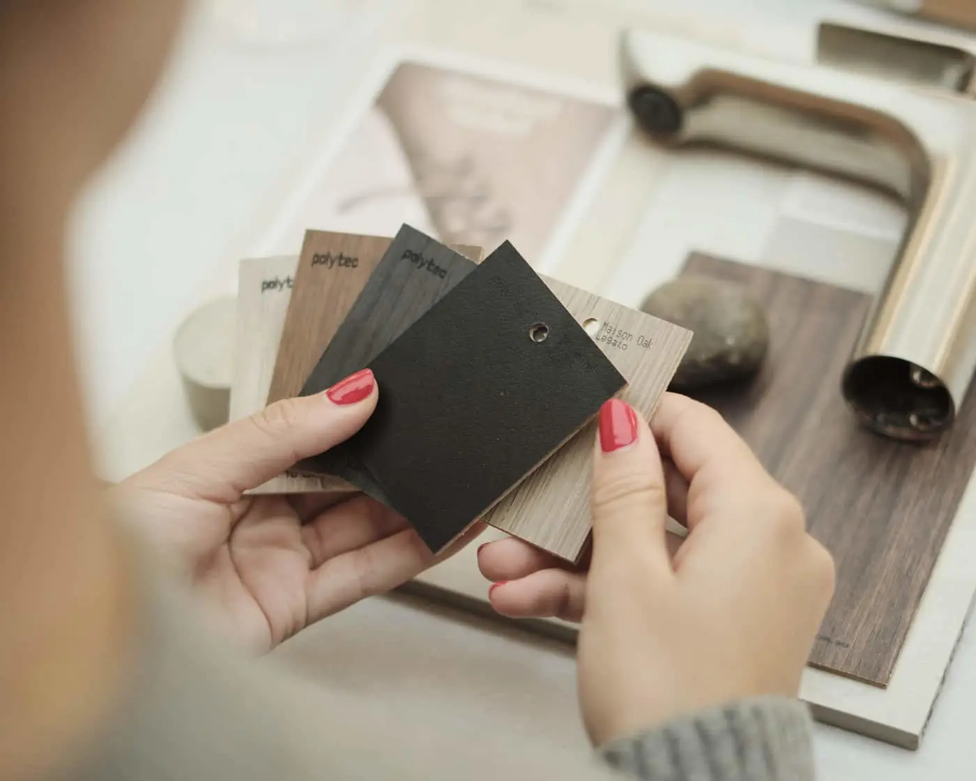 A person with red nails holds several Polytec material swatches in wood and dark tones, with cabinet hardware samples in the background.