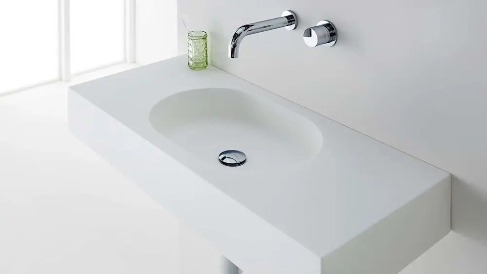 Modern white bathroom sink with wall-mounted chrome faucet and small green textured glass container nearby.