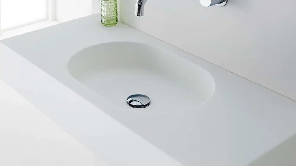 Modern white bathroom sink with wall-mounted chrome faucet and small green textured glass container nearby.