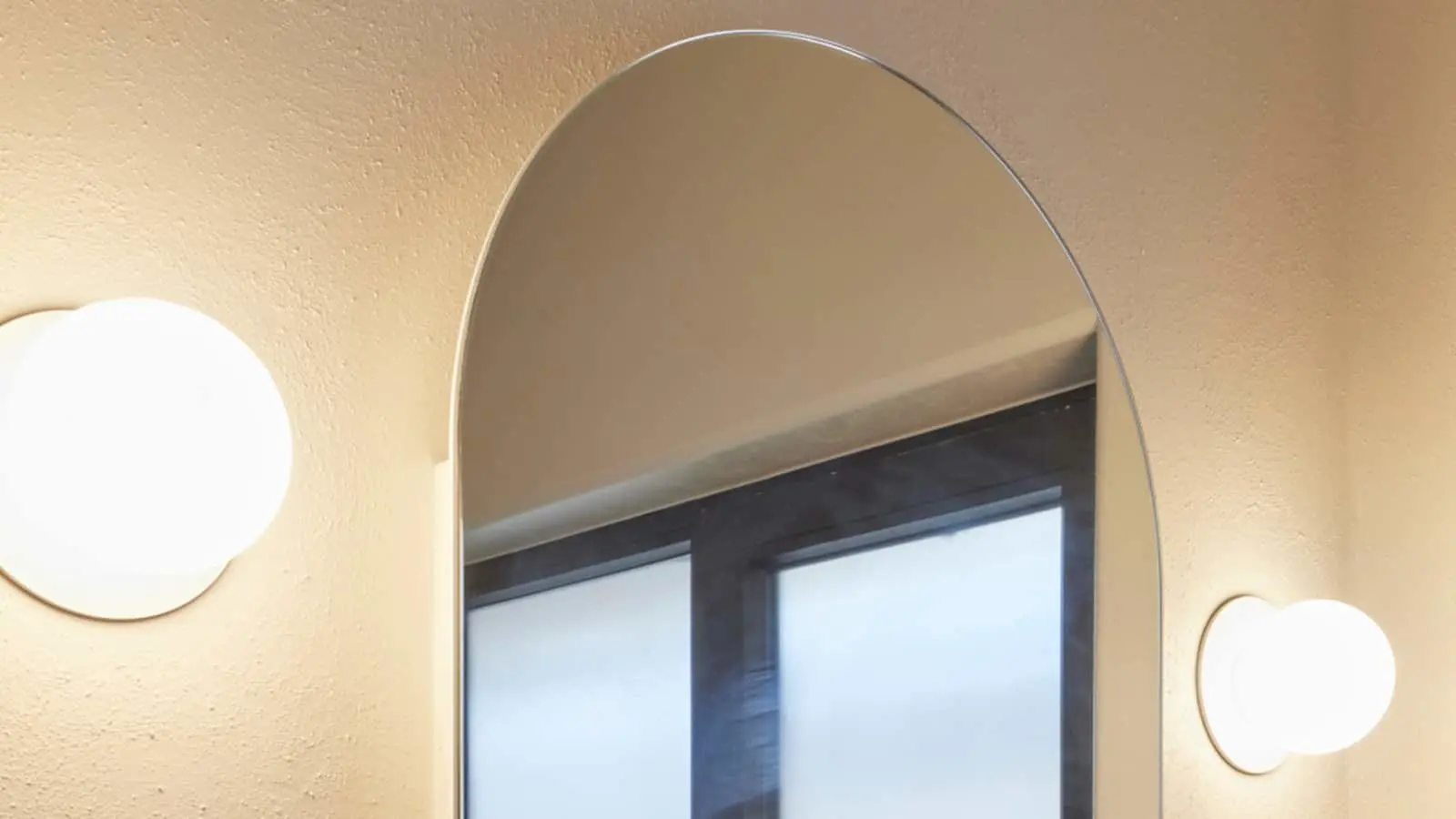 Oval frameless mirror on beige wall with round wall lights on either side, reflecting a window.