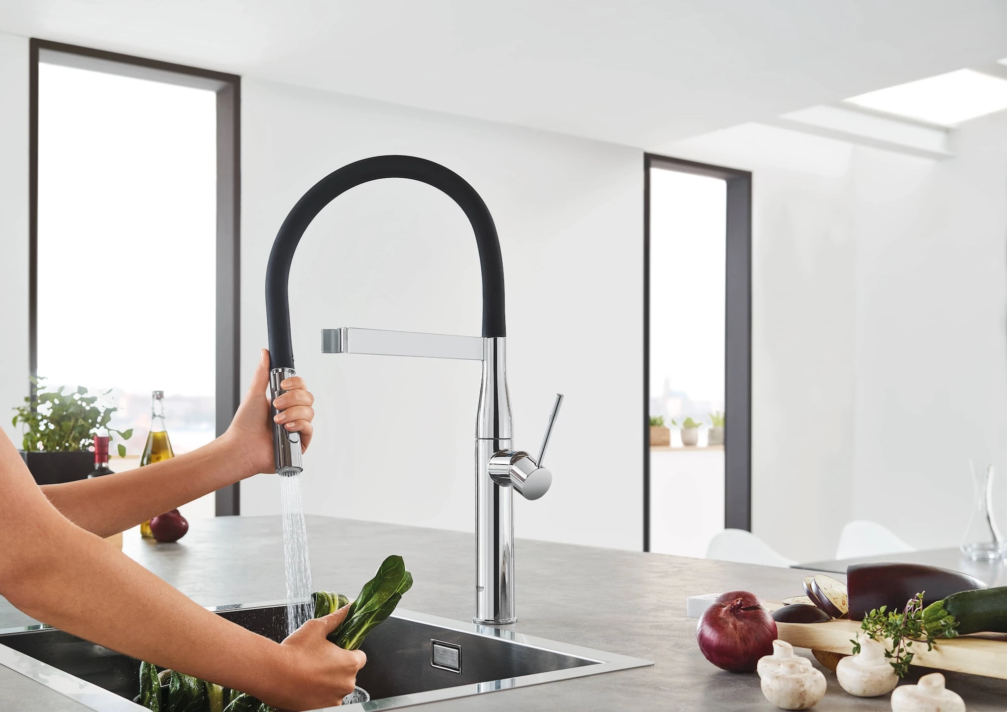 Hand using modern kitchen faucet with flexible black hose to rinse vegetables in sink, fresh produce on counter.