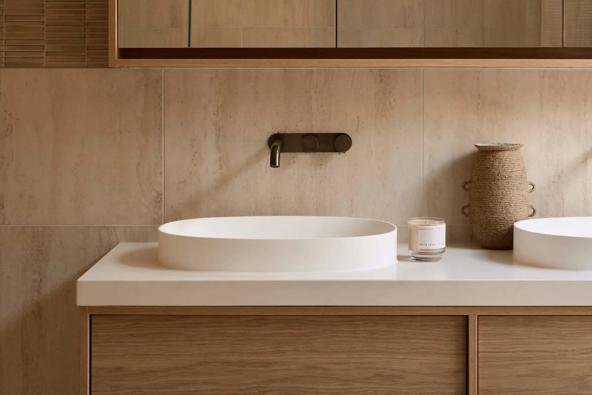 Bathroom vanity with oval white basin, dark wall-mounted tap, candle, woven vase, wood cabinetry, and beige stone tiles.