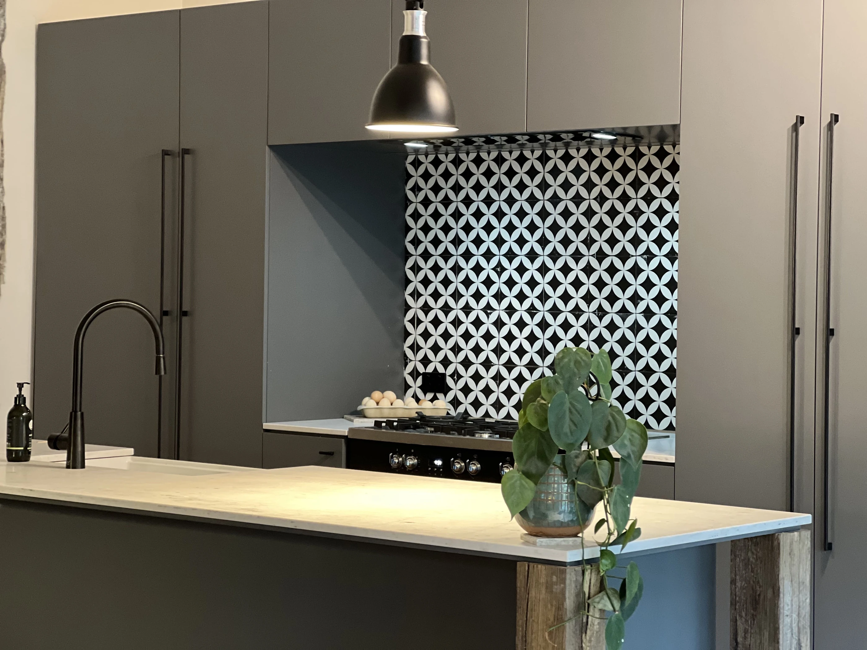 Modern kitchen with gray cabinets, black and white patterned backsplash, pendant light, and potted plant on counter.
