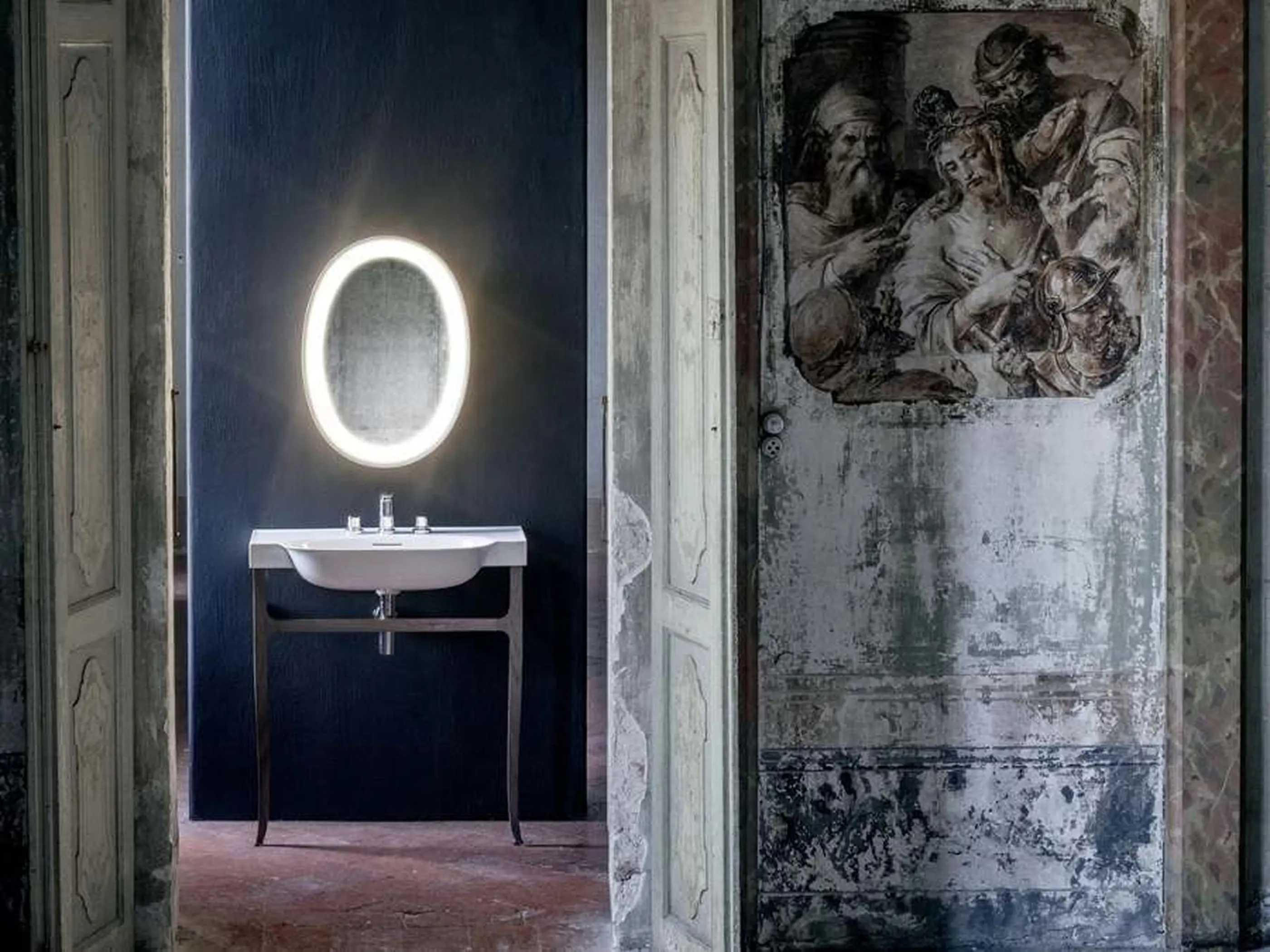 Modern bathroom with illuminated round mirror and white sink contrasting with historic wall featuring a classical fresco painting.