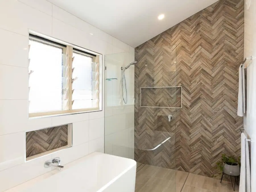 Modern bathroom with white tub, glass shower, and feature wall of wooden herringbone tiles beside white walls.